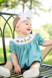 Baby girl sitting on porch swing wearing Smocked Camping Bubble by Ruth and Ralph, teal gingham