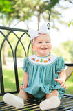 Baby girl sitting on porch swing wearing Smocked Camping Bubble by Ruth and Ralph, white bow, outdoors