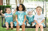 Toddler sitting on porch swing wearing Smocked Camping Jon Jon by Ruth and Ralph, holding a ball
