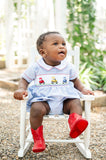 Baby boy standing in white chair wearing Smocked Tractor Bubble by Ruth and Ralph with red boots