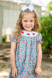 Girl standing outdoors in Ruth and Ralph Smocked Floral Pumpkin Dress, blue floral print with white bow