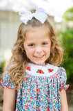 Girl smiling in Smocked Floral Pumpkin Dress by Ruth and Ralph, blue floral print with white bow, outdoors
