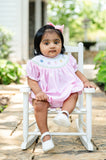 Baby girl sitting in white chair outdoors wearing Smocked Rainbow Pumpkins Dress by Ruth and Ralph