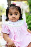 Baby girl wearing Smocked Rainbow Pumpkins Dress by Ruth and Ralph, pink with white bow, sitting in white chair outdoors