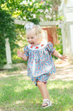 Baby girl running and laughing in Smocked Floral Pumpkin Bloomer Set by Ruth and Ralph, blue floral print with white bow, outdoors