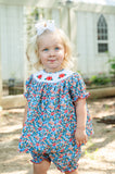 Baby girl standing outdoors wearing Smocked Floral Pumpkin Bloomer Set by Ruth and Ralph, blue floral print with white bow