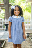 Girl standing outdoors in Ruth and Ralph Smocked Hunting Season Dress, blue gingham with white bow