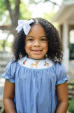 Girl smiling in Smocked Hunting Season Dress by Ruth and Ralph, blue gingham with white bow, outdoors