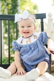 Baby girl wearing Smocked Hunting Season Bubble by Ruth and Ralph, blue gingham with white bow, sitting outdoors