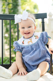 Close-up of baby girl smiling in Smocked Hunting Season Bubble by Ruth and Ralph, sitting outdoors with white bow