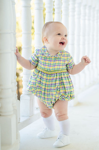 A baby in a green, yellow, and purple plaid smocked Mardi Gras bubble with intricate details.