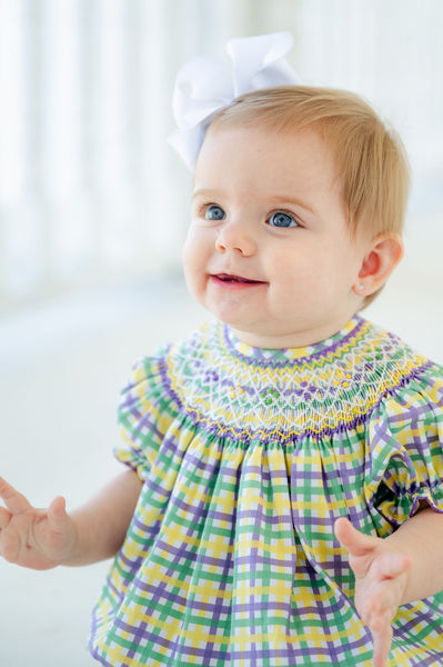 A baby girl in a smocked Mardi Gras bubble looks festive and adorable in the vibrant green, yellow, and purple plaid.