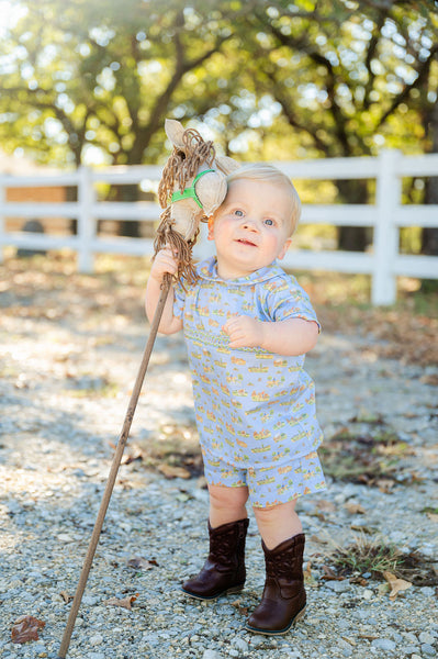 A little boy wearing the Smocked Cowboy Toile Samuel Shirt/Shorts Set looks adorable in this charming outfit featuring a cowboy toile print on a blue background.