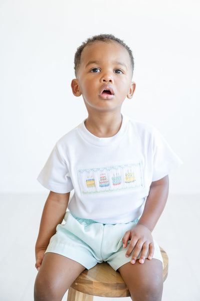 A little boy beams in his smocked birthday cake T-shirt and mint green shorts, ready for his big celebration.