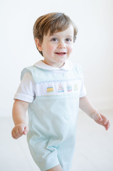 A little boy beams in his smocked mint green jonjon, featuring birthday cake details, perfect for his big day.