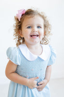 The Classic Blue Smocked Dress for girls features a hand-smocked bodice adorned with delicate light blue and pink flowers.