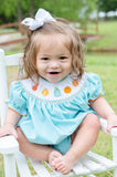 Baby girl smiling in Smocked Fall Gourds Bubble, teal gingham with white bow, sitting on white chair outdoors
