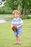 Toddler boy wearing a blue gingham smocked football jonjon with three hand-smocked footballs over a Peter Pan collar shirt, holding a football