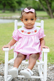 Baby girl wearing a pink gingham smocked football bubble with a hand-smocked neckline of footballs and bows, sitting in a white rocking chair