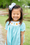 Girl smiling in Smocked Fall Gourds Dress by Ruth and Ralph, teal gingham with white bow, outdoors