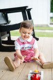 Baby boy sitting on porch playing with toy wagon, wearing Smocked Race Car Jon Jon by Ruth and Ralph, red gingham
