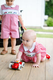 Two children playing outdoors, baby crawling on porch wearing Smocked Race Car Bubble by Ruth and Ralph, red gingham
