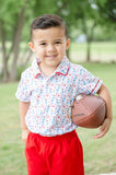 Toddler boy wearing a white Pima cotton polo with football and tailgate print, red piping, and solid red shorts, holding a football