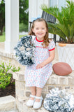 Toddler girl wearing a white Pima cotton football dress with game day print and red trim, holding silver pom-poms