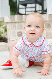Smiling baby in a white and red game day bubble with football and tailgate motif print, sitting on a brick step